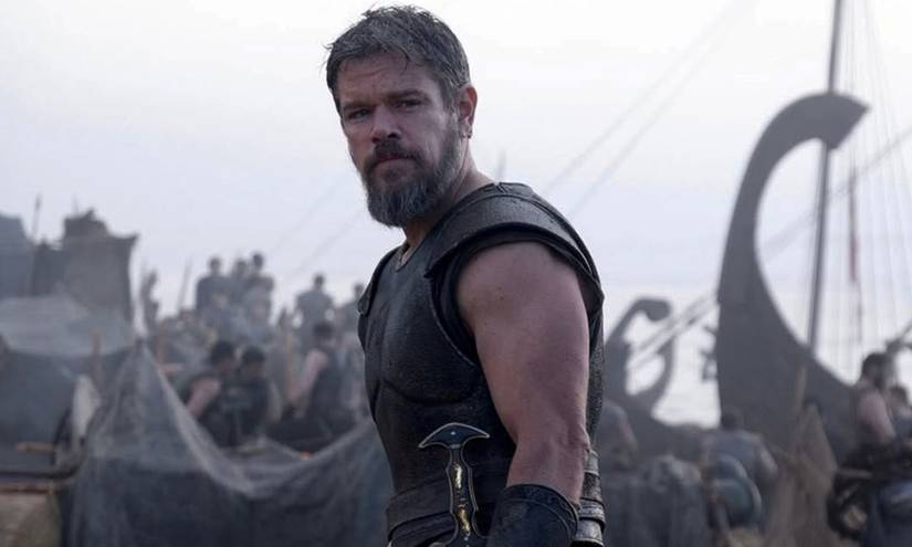 Matt Damon, bearded and armored as Odysseus, stands on the deck of a Greek ship with other soldiers and curved prows in the background in Christopher Nolan’s The Odyssey.