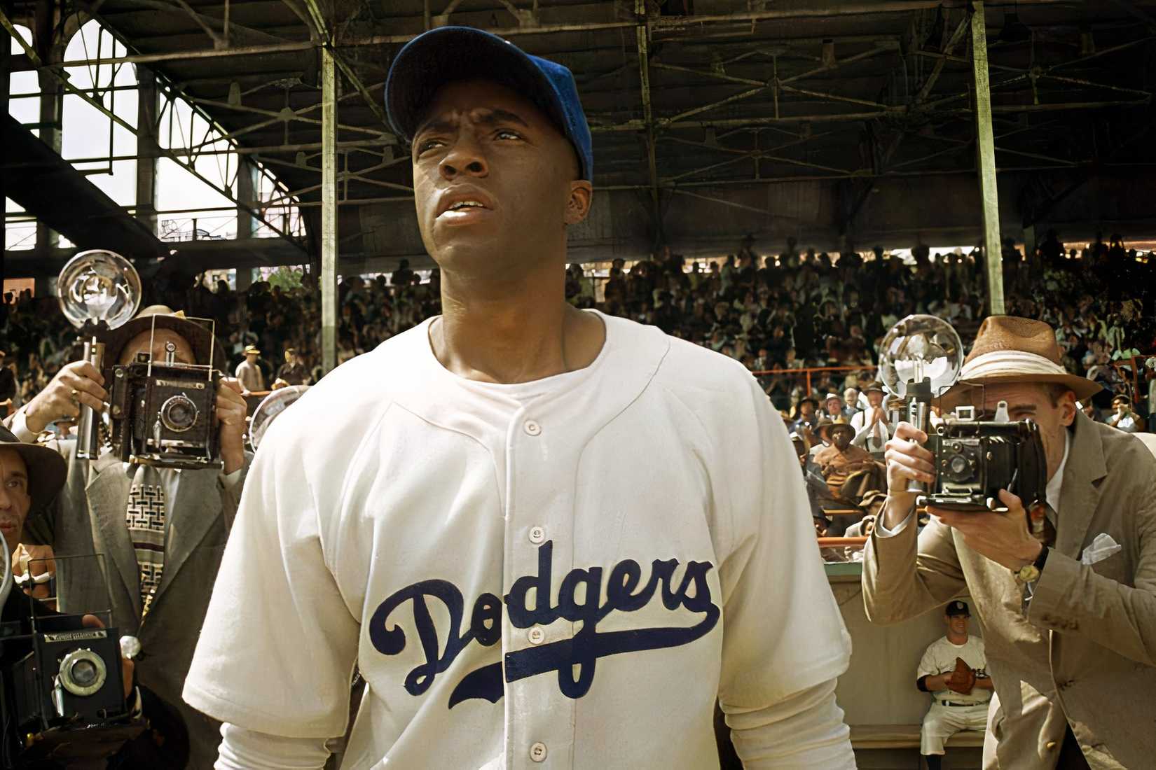 Jackie Robinson stands on a baseball field in a Dodgers uniform as photographers crowd around him in 42.