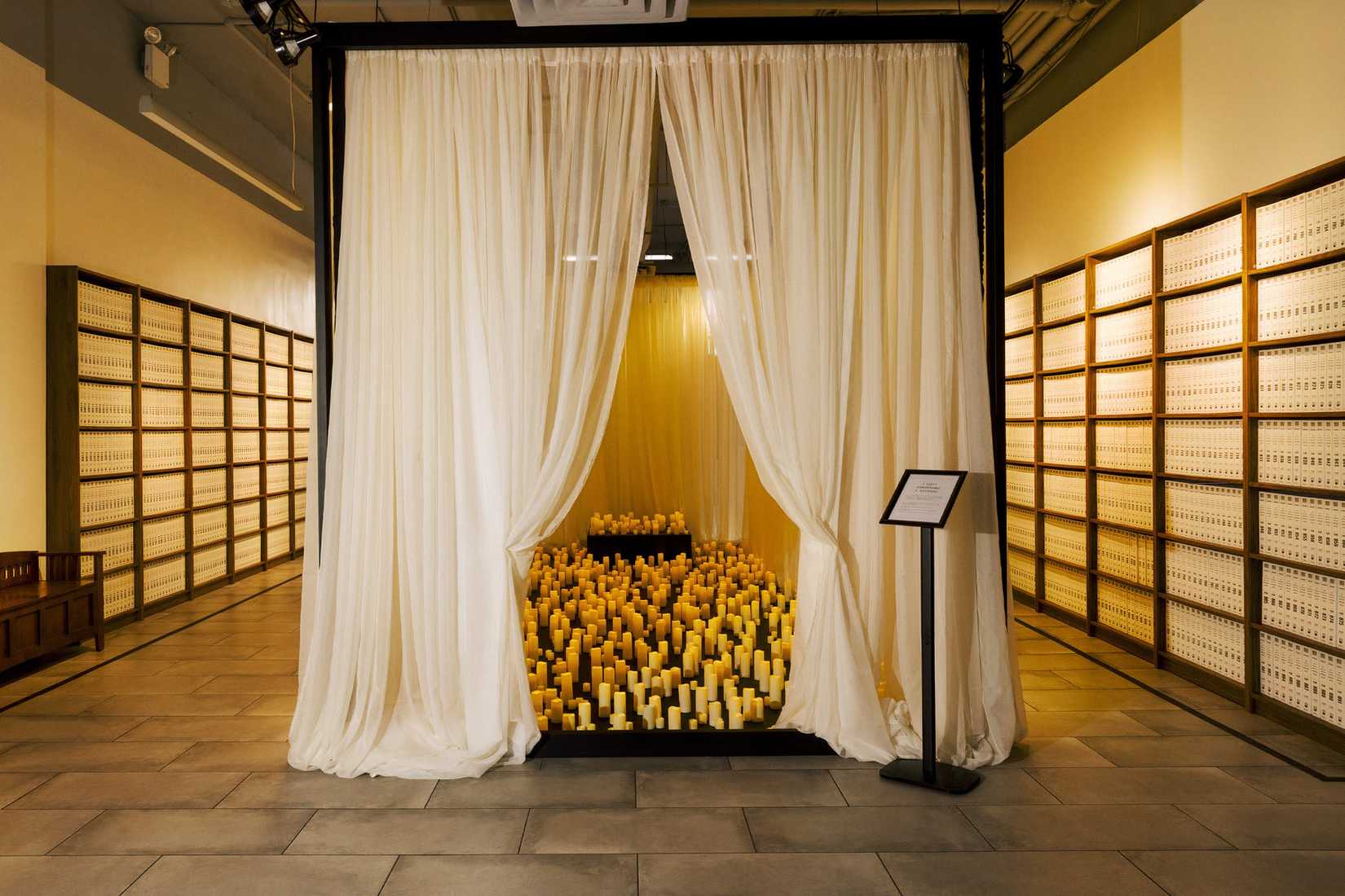 A wide view of the candlelit tribute area, with shelves of bound Epstein files lining the room on both sides.
