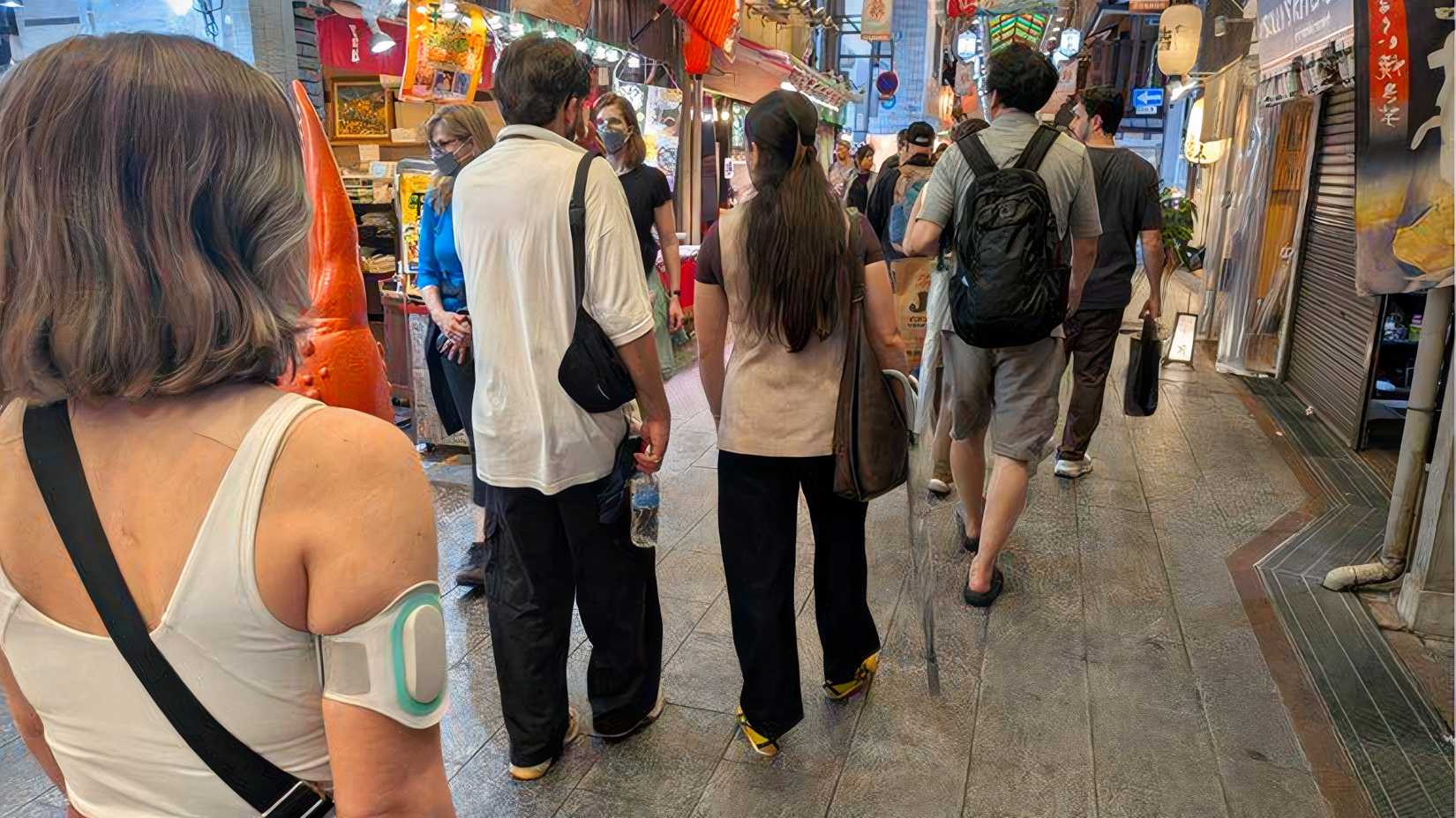 TJ walks through a crowded indoor market in Japan with a Nerivio migraine device visible on her upper arm.