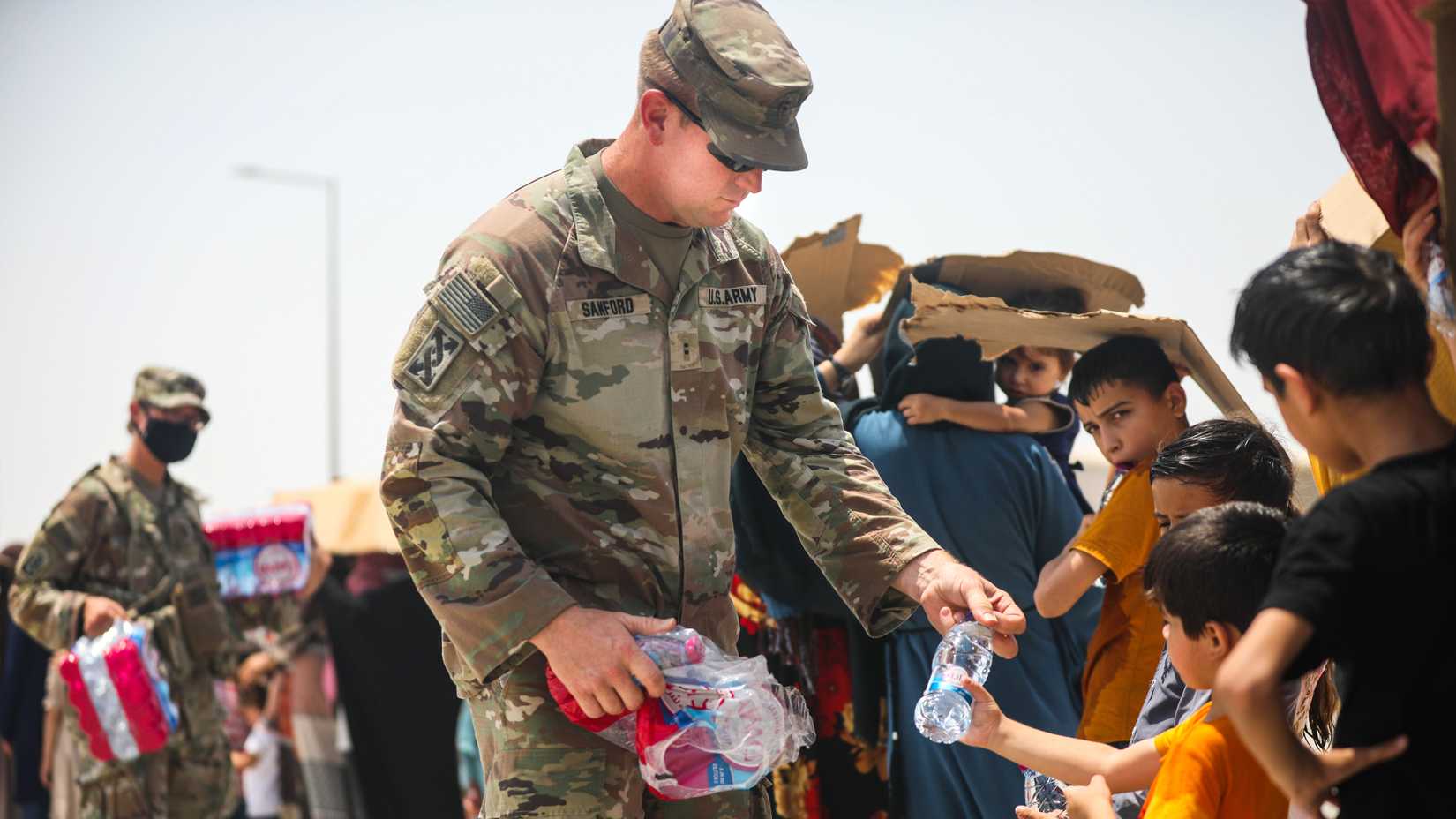 U.S. Army soldier in camouflage hands bottled water to Afghan children during the 2021 evacuation from Afghanistan.