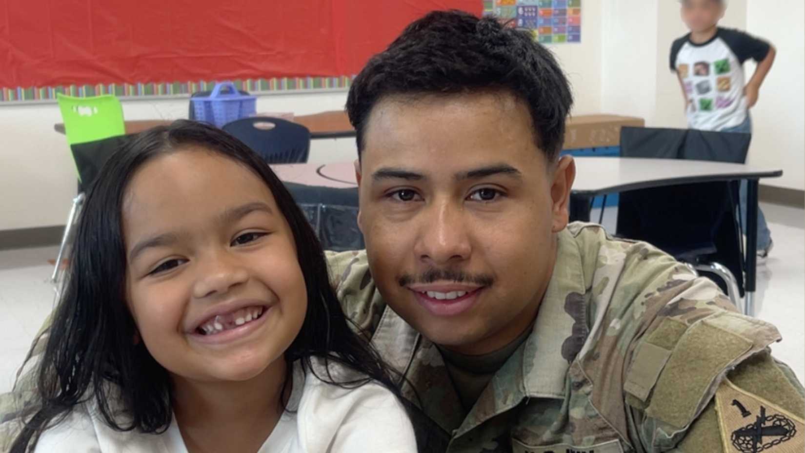 Army Sgt. Hedár Leonel Turcios Juárez, wearing his camouflage uniform, smiles beside his young daughter in a classroom.