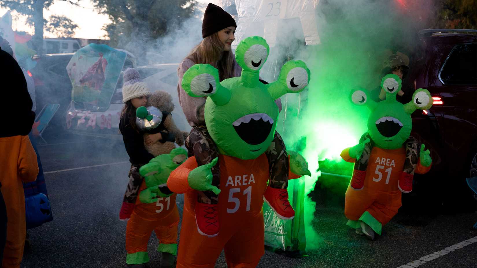 A military family wears matching green alien Area 51 costumes beside a decorated vehicle at a Halloween event.