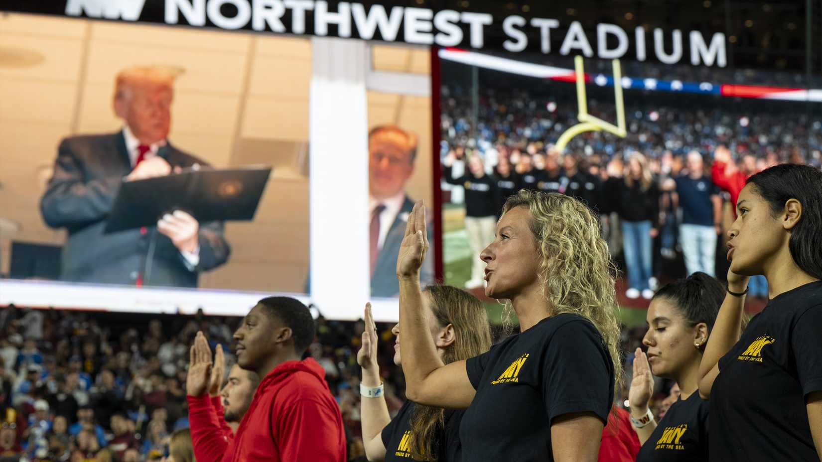 Military recruits raise their right hands for a swearing-in ceremony as President Donald Trump appears on a stadium video screen during a Washington Commanders game.