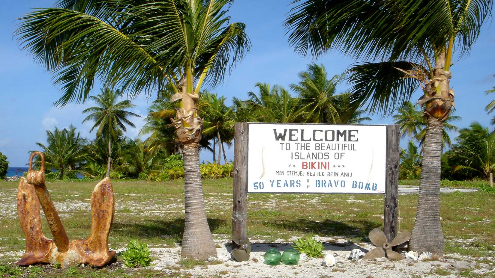 A welcome sign on Bikini Atoll stands among palm trees and rusted naval artifacts, marking 50 years since the Castle Bravo nuclear test.