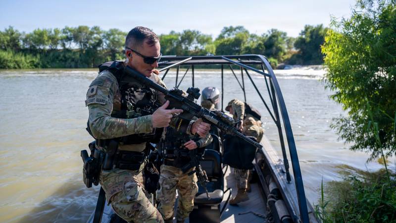Armed personnel in tactical gear ride a patrol boat on a river