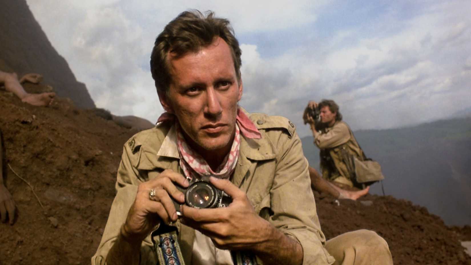 James Woods portraying photojournalist Richard Boyle holding a camera while crouching outdoors in a scene from Salvador (1986).